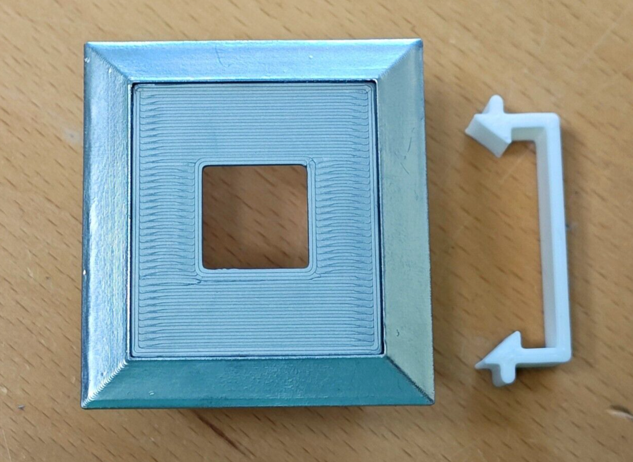 Silver right power window switch bezel with a central square cutout, shown next to a white plastic retainer clip. Compatible with 1980-1986 Ford F-Series trucks and Bronco.