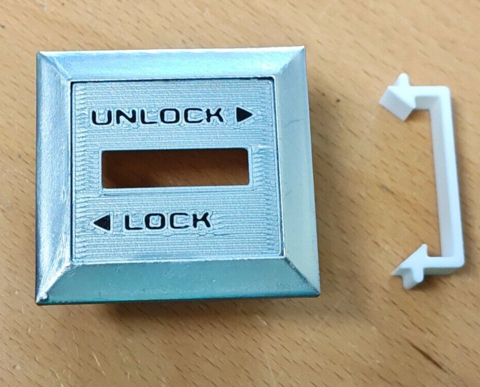 Silver reproduction left power lock bezel with black UNLOCK and LOCK text and arrows, shown next to its white plastic retainer clip for 1980-1986 Ford F-150, F-250, F-350, and Bronco.