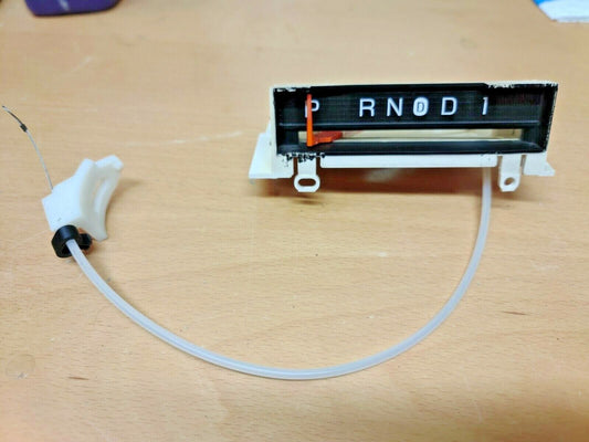 Ford 1980-1986 automatic transmission shift indicator for AOD overdrive. Features black face with P, R, N, circled D, D, 1 lettering, red needle pointer, and attached adjustment cable.