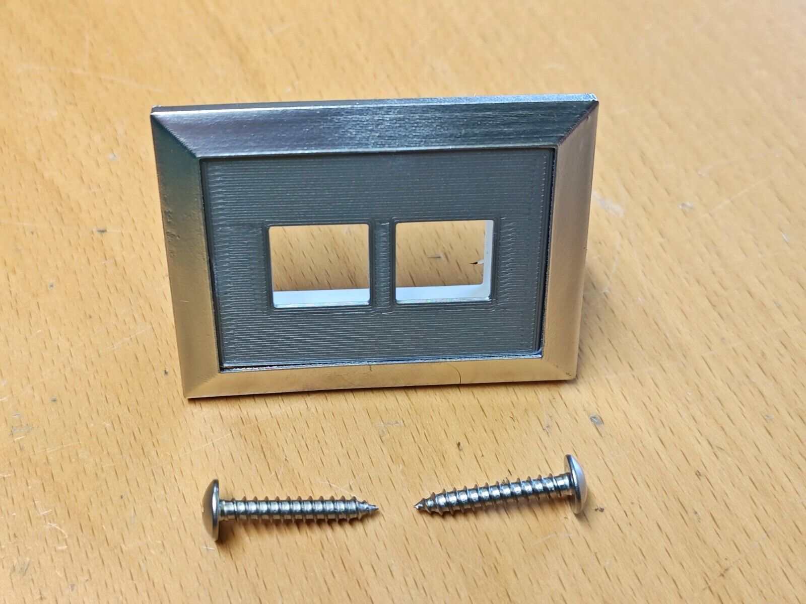 Left power window bezel with a chrome frame, textured gray center, and two square switch openings, shown alongside two silver mounting screws. Compatible with 1980-1986 Ford F-150, F-250, F-350, and Bronco models.
