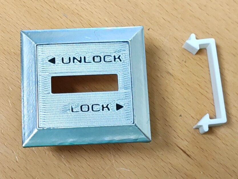 Silver passenger side power door lock switch bezel with black UNLOCK and LOCK text, shown next to a white plastic retainer clip. For 1980-1986 Ford F-Series trucks and Bronco.